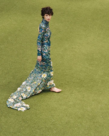 Woman in a floral gown standing on green grass, photographed outdoors. She is captured in a high angle shot, looking at the camera.
