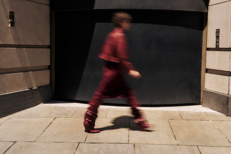 Person in red outfit walks on a city sidewalk past a dark doorway. The image is intentionally blurred, creating a sense of movement.