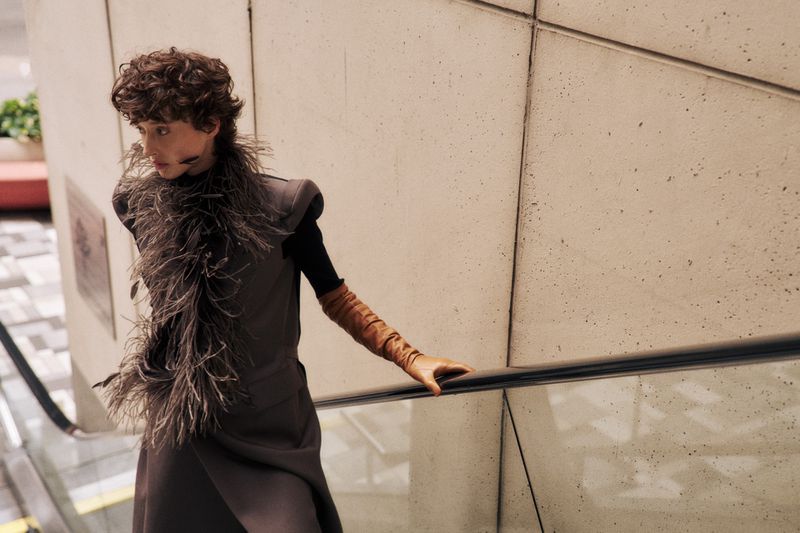 Fashionable woman in grey coat and brown gloves poses on escalator with neutral tiled walls, creating a stylish editorial image.