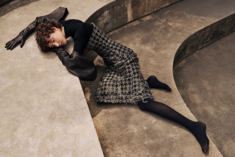 Fashion shot of a young person in a black turtleneck and houndstooth skirt lying on a concrete surface, wearing oversized black gloves.