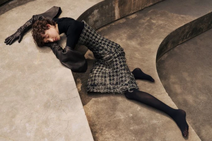 Fashion shot of a young person in a black turtleneck and houndstooth skirt lying on a concrete surface, wearing oversized black gloves.