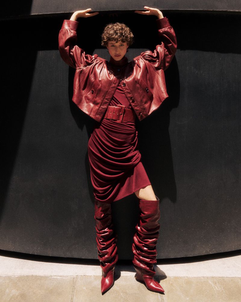 A woman in a burgundy leather jacket, dress, and thigh-high boots poses with her arms up against a dark background in sunlight.