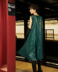 Model Madeleine German in a green pleated dress and black opera gloves standing on a subway platform by a Broad St pillar sign.