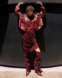 Model posing with arms raised under an alcove wearing a monochrome burgundy leather jacket, draped dress, and slouchy high boots.