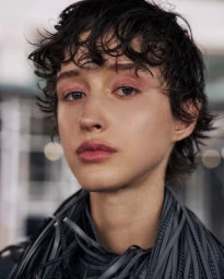 Close-up portrait of model Madeleine German with wet-look curly dark hair and pink eye makeup wearing a grey fringed collar.