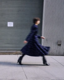 Motion blurred model wearing a dark long coat walking rapidly along a city sidewalk next to a corrugated metal wall.