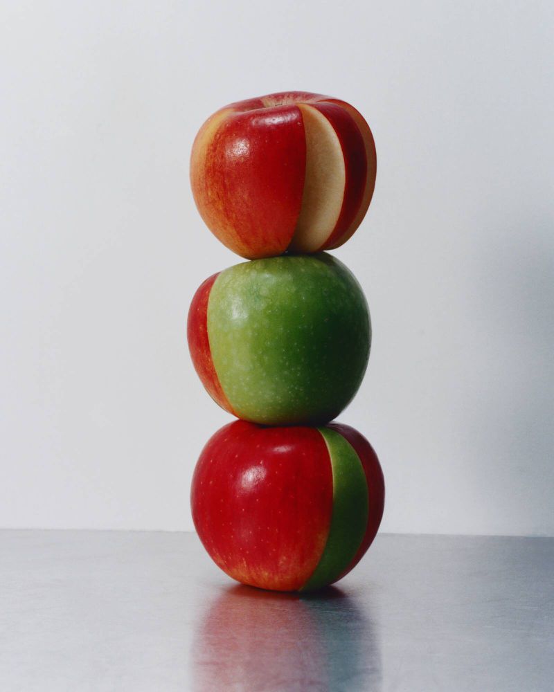 Stacked red and green apples create a colorful, balanced fruit still life on a reflective surface in a studio setting.