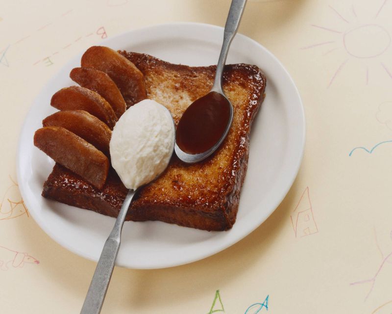 Overhead shot of French toast with caramelized apples, cream, and chocolate syrup, served on a white plate with a patterned background.