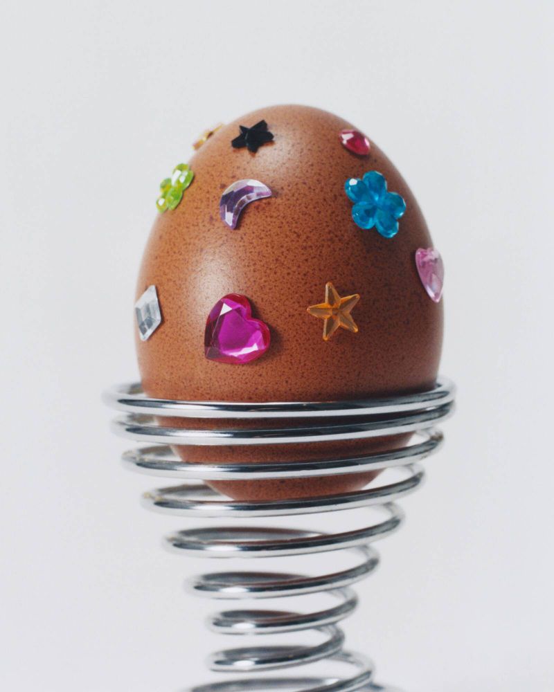 A brown egg decorated with colorful sparkly shapes sits in a silver coiled wire egg cup against a solid white background. Food still life.