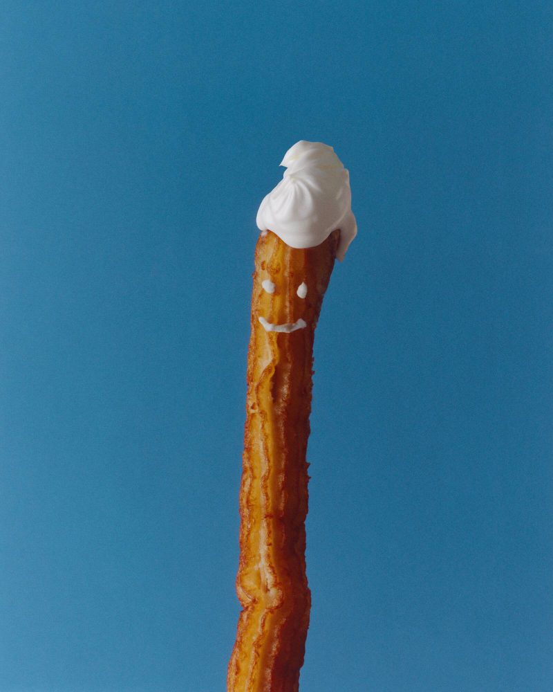 Close-up of a churro standing upright against a blue background, topped with a dollop of white cream and a smiling face drawn on the churro.