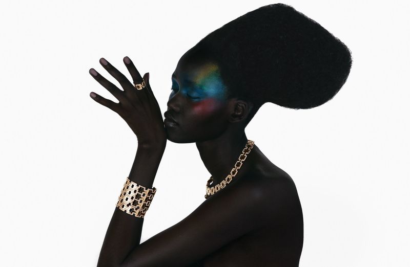 Model Caren Jepkemei wearing gold jewellery and colourful eye makeup with a sculpted bouffant hairstyle against a white background.