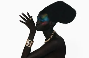Model Caren Jepkemei wearing gold jewellery and colourful eye makeup with a sculpted bouffant hairstyle against a white background.