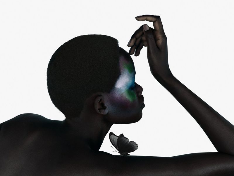 Striking beauty portrait of a woman with dramatic green and purple makeup, complimented by a black butterfly on her shoulder against a white background.