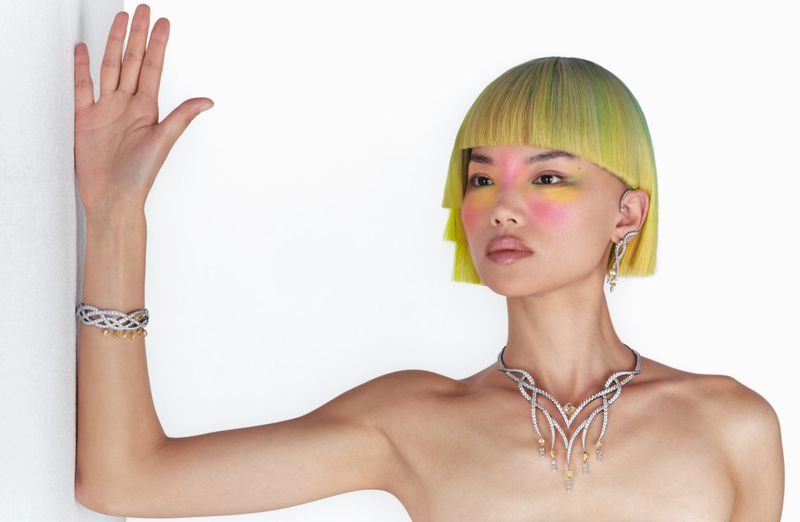 Woman with colorful hair and makeup wearing earrings, necklace, and bracelet jewelry in a fashion portrait against a white background.