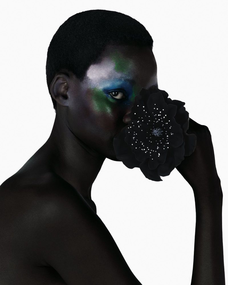 Striking portrait of a Black woman with vibrant blue and green makeup, holding a large black flower against a clean white background.