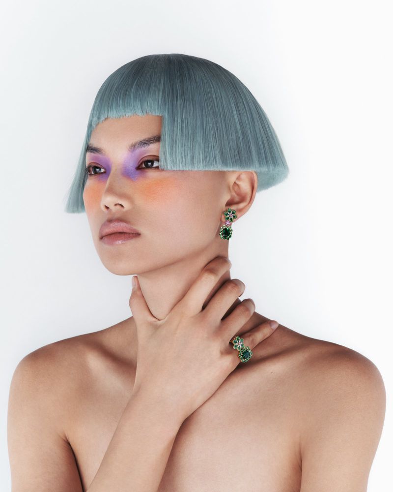 Asian woman with bold makeup, blue hair, and jewelry in a studio shot on a white background showcasing fashion and beauty elements.