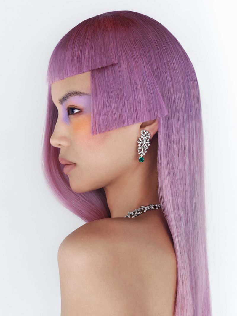 Profile portrait of a woman with short purple hair, striking makeup, and statement jewelry, set against a clean white backdrop.