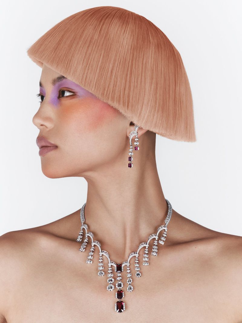 Model showcases diamond necklace and earrings with a short blond bob hairstyle, dramatic eye and cheek makeup in a studio shot on white.