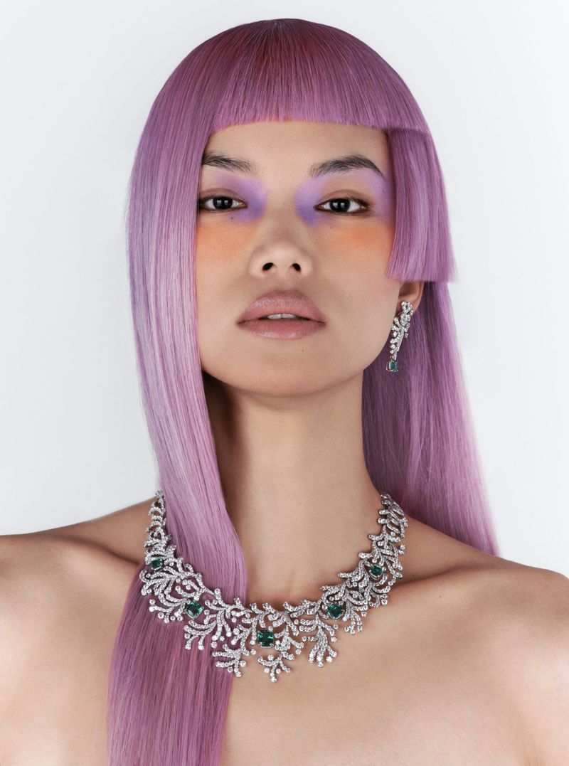 Close-up studio shot of an Asian female model with purple blunt-cut hair, vibrant eye makeup, and a floral-inspired diamond necklace and earrings.
