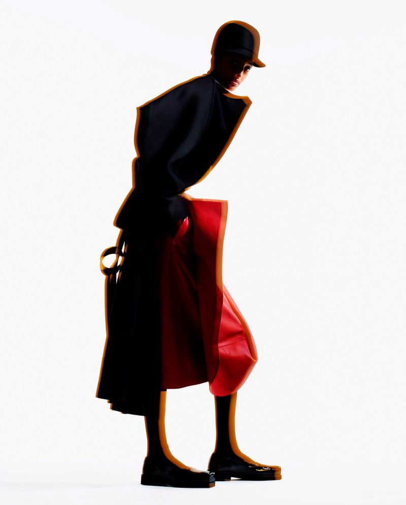 Fashion portrait of adult woman in black hat and coat with red skirt, posing against a white backdrop in a photo studio setting.
