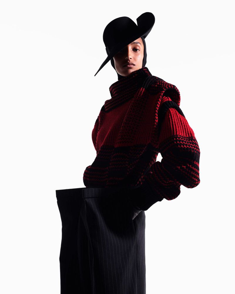 Fashion-forward woman in black headwear and oversized red/black knitwear, captured in a high-key studio shot with clean lines and a modern aesthetic.