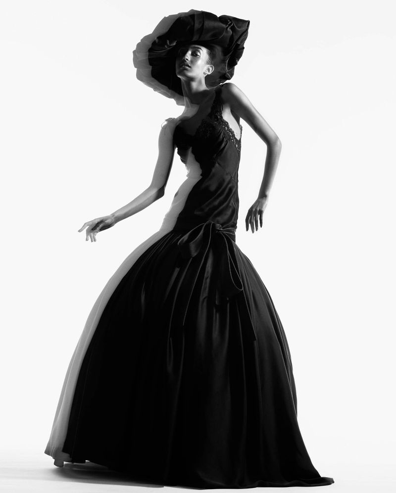 Black-and-white portrait of a fashion model wearing a dramatic black dress and hat, set against a bright white background.
