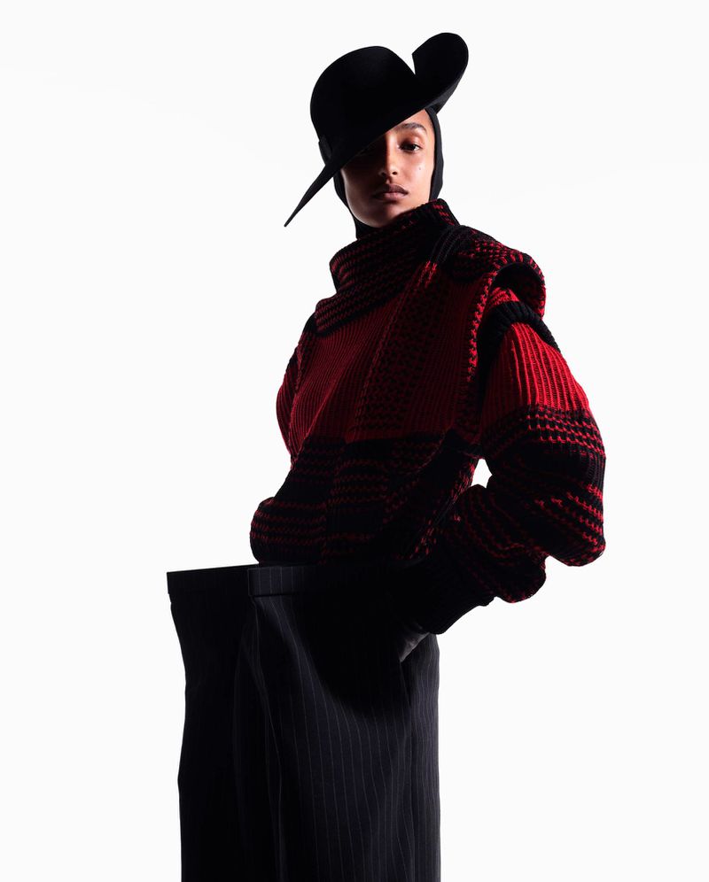 Fashion shot of a female model in a red and black sweater with a unique black hat, posing in a photo studio against a white backdrop.