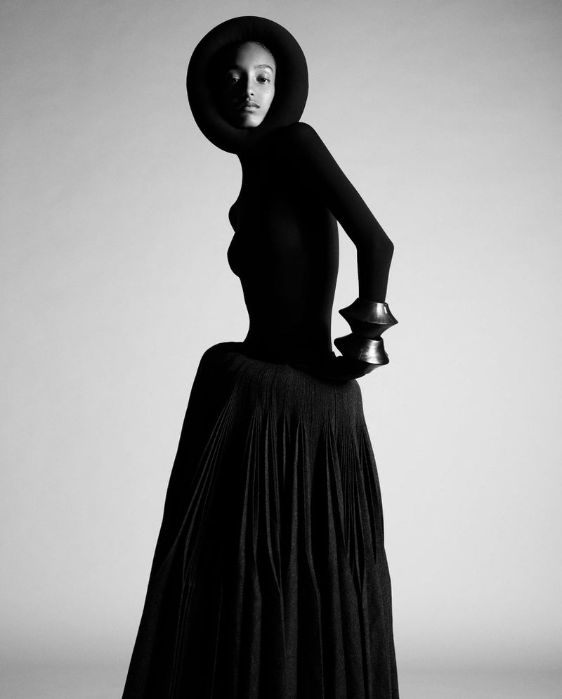 A black-and-white fashion portrait of a woman in an avant-garde black dress with sculptural sleeves and a circular headpiece.