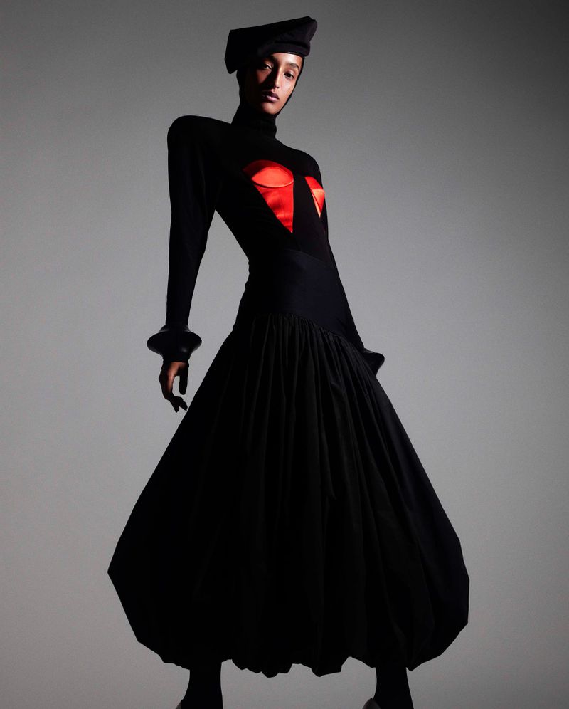 Woman in black gown, cap, and striking red detail poses elegantly against a neutral backdrop in a studio fashion photograph.