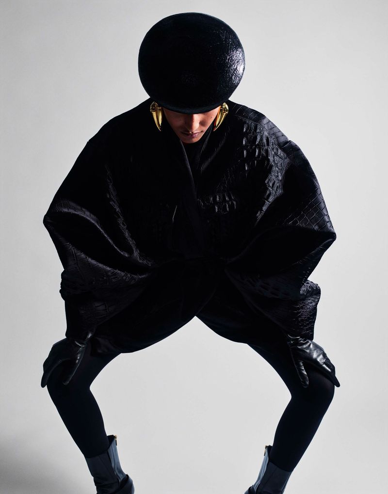 Model Mona Tougaard crouches in a voluminous black textured cape, rounded black hat, and gold earrings against a white seamless backdrop.