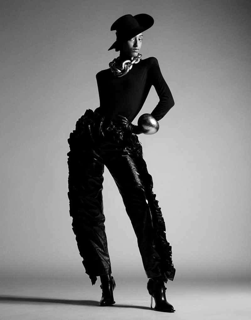 Black and white studio portrait of a slender fashion model wearing hat, jewelry, body suit, and ruffle pants, posing against a plain backdrop.