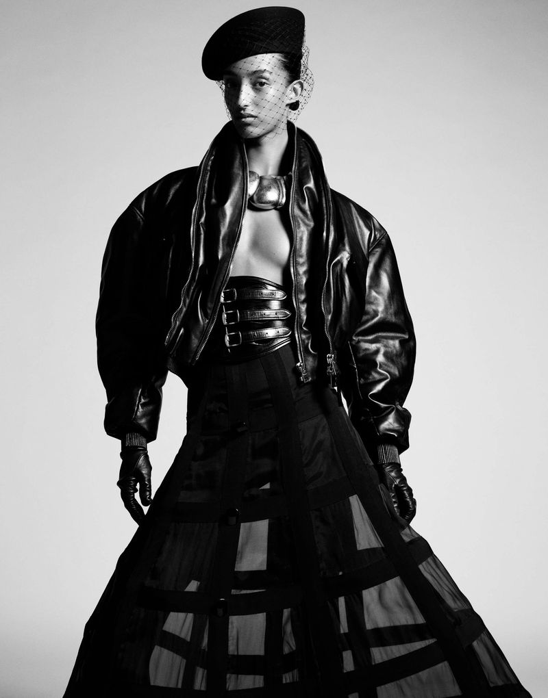 Black and white fashion portrait of a woman wearing a beret, oversized leather jacket, wide belt, and sheer skirt in a studio.