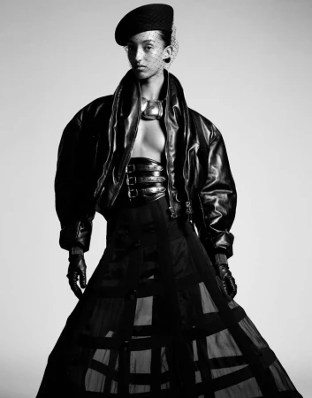 Black and white fashion portrait of a woman wearing a beret, oversized leather jacket, wide belt, and sheer skirt in a studio.