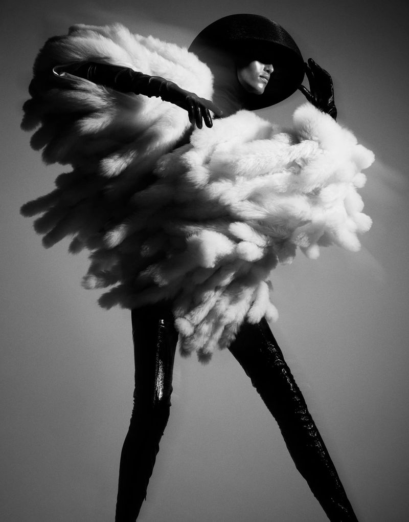 Striking black and white fashion shot of a model wearing a large, fluffy white coat, hat, and shiny black leggings against a gray backdrop.