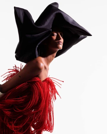 Model Mona Tougaard wearing an oversized sculptural black hat and textured red fringe garment against a white background.