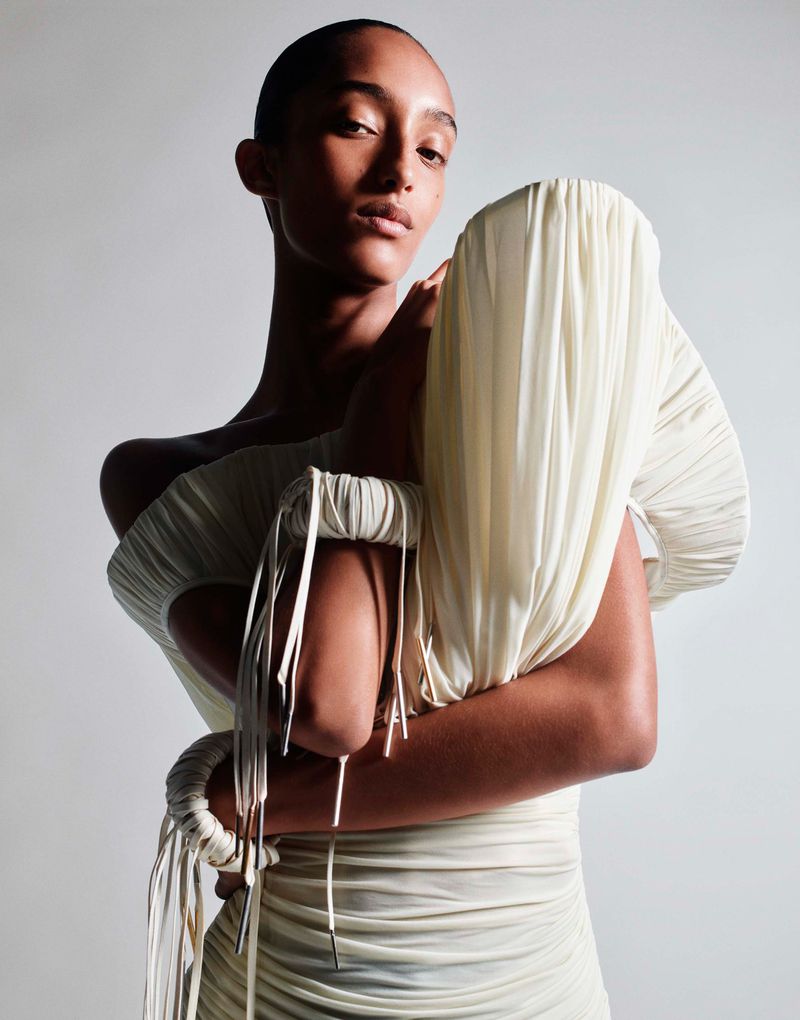 A solo female model poses in a studio wearing a light cream, form-fitting dress, with folded textures and tied elements on the upper arms.