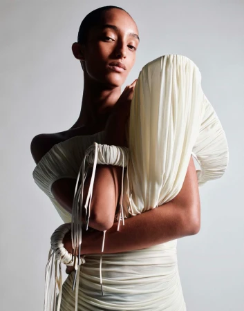 A solo female model poses in a studio wearing a light cream, form-fitting dress, with folded textures and tied elements on the upper arms.