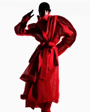 Model Mona Tougaard posing in a sculptural red belted coat with voluminous sleeves and a black cap against a stark white background.