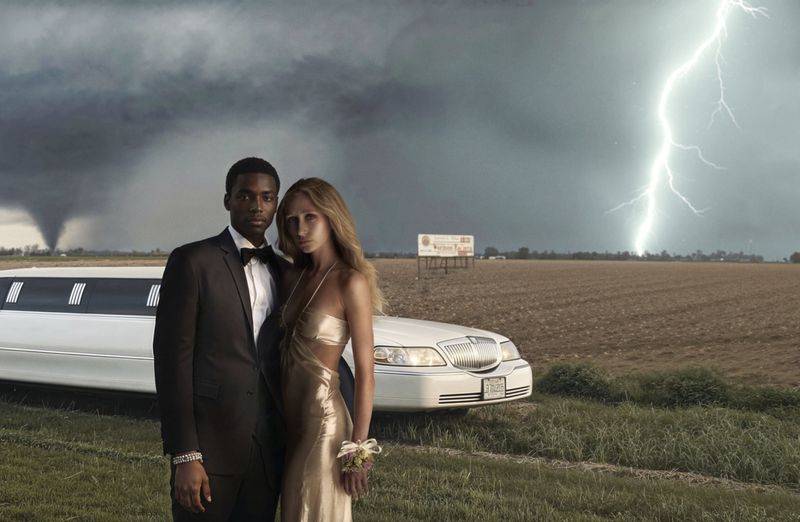 A couple in formalwear poses in a field near a white limo during a lightning storm with a tornado in the distance. 