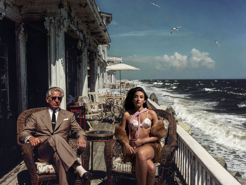 Man and woman in swimwear sit on a porch overlooking the ocean with seagulls and waves, bathed in sunny daylight.