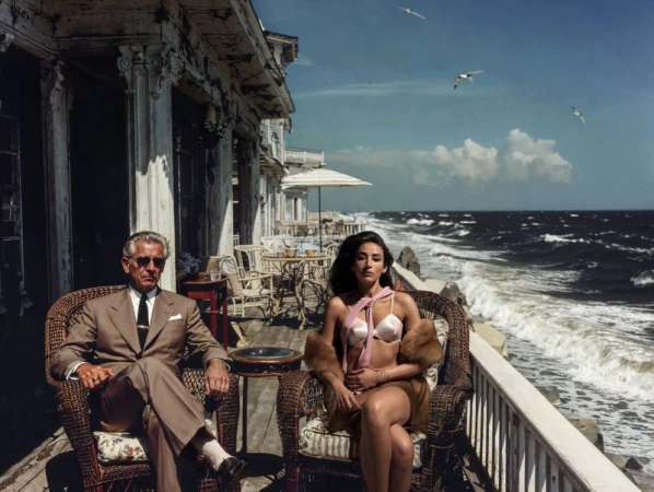 Man and woman in swimwear sit on a porch overlooking the ocean with seagulls and waves, bathed in sunny daylight.