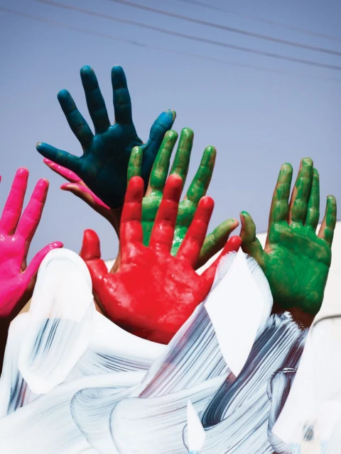 Colorful hands raised against a bright sky, covered in vibrant red, green, teal, and pink paint, creating a cheerful, artistic display.