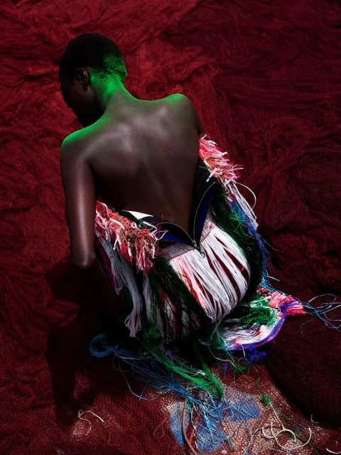 Back view of dark-skinned woman in fringed dress, spotlight on hair, red textured background. Fashion, beauty, and creative photography.