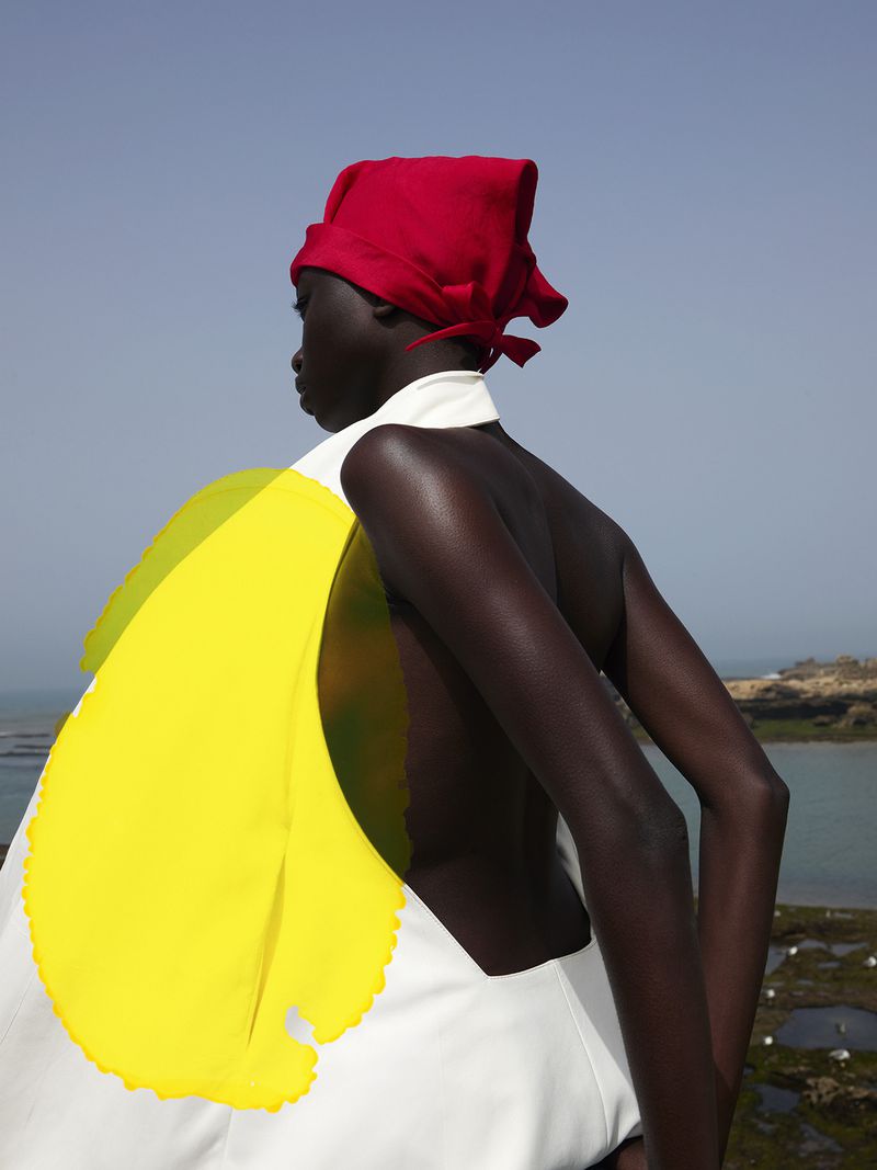 Woman in a red headscarf, yellow shawl and white dress stands outdoors with a sea view on a clear day, fashion editorial.