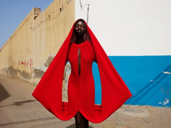 A dark-skinned woman in a bright red dress with wide, cape-like sleeves stands in an alleyway, posing against a weathered wall on a sunny day.