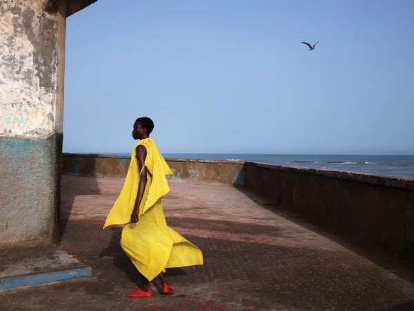 A woman in a yellow outfit walks along a concrete structure near the ocean under a blue sky with a bird flying above.