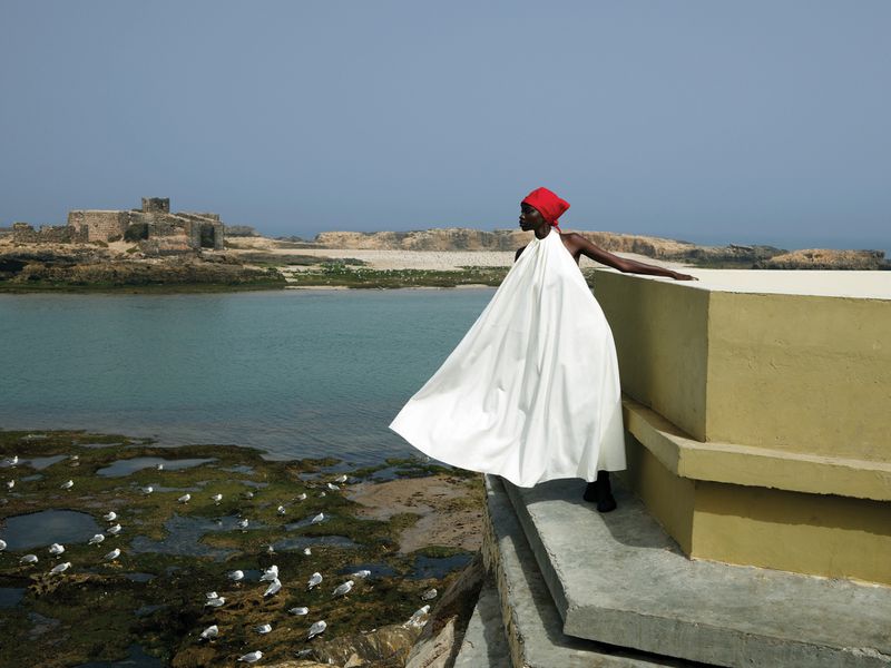 A woman stands on a ledge in a draped white gown with a red head wrap, looking out over the sea on a bright, sunny day.