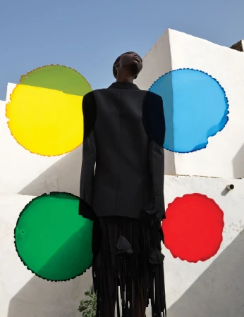 Fashion shot of a Black woman in a black suit against a white wall with colorful circles, looking up at a clear blue sky.