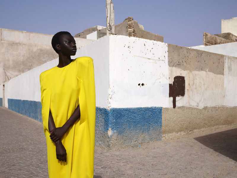 An African American woman poses in a yellow dress on a cobblestone street, against a backdrop of weathered white and blue buildings.