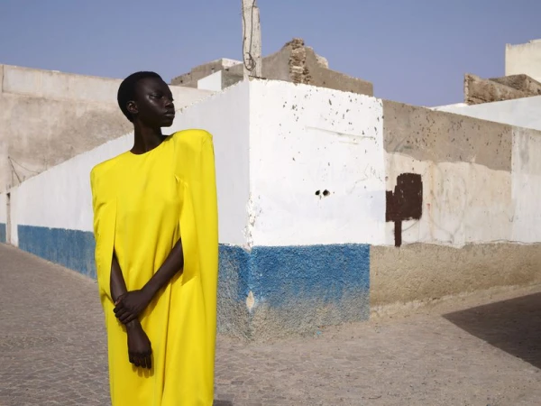 An African American woman poses in a yellow dress on a cobblestone street, against a backdrop of weathered white and blue buildings.